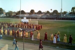 Students in academic regalia participating in a graduation ceremony on a sports field, with spectators and a track in the background.